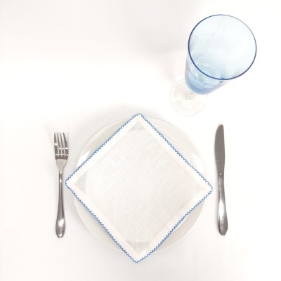 Conjunto de mesa com prato branco, guardanapo branco com bordado azul, talheres de metal e copo azul.
