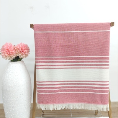 Manta ou toalha rosa com riscas brancas pendurada num suporte de madeira com vaso branco e flores cor-de-rosa ao lado