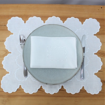 Conjunto de prato cinza com guardanapo branco bordado, talheres inox, e base de tecido branco bordado em mesa de madeira.