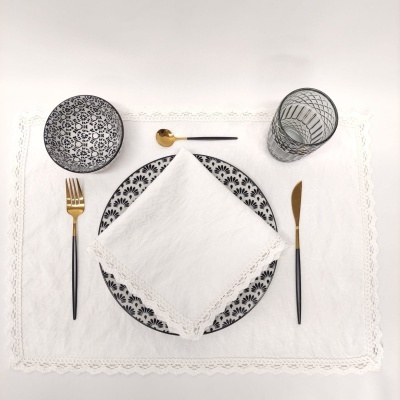 Conjunto de loiça preta e branca com padrão floral, talheres dourados, guardanapo branco com renda, tudo sobre base branca rendada