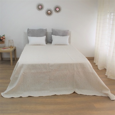 Cama de casal com colcha branca e almofadas cinzentas e brancas perto de mesa de cabeceira com flores e lâmpada.