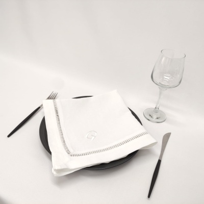 Conjunto de loiça com prato preto, guardanapo branco bordado, talheres com cabo preto e copo de vinho em mesa branca