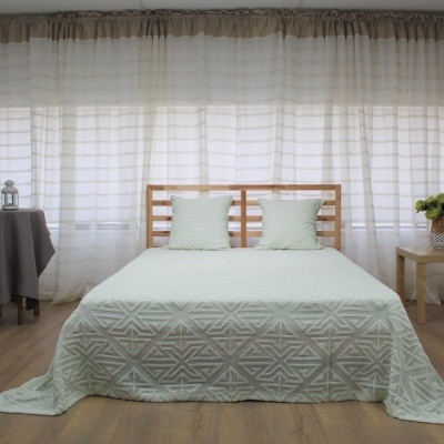 Cama de madeira clara com colcha verde clara e almofadas, mesas laterais com decoração e cortinas brancas