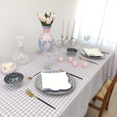 Mesa de jantar com toalha xadrez lilás e branca, pratos brancos com padrão azul, copos, talheres dourados, castiçais com velas cor-de-rosa, flores em vaso rosa com padrão azul e garrafa de vidro trabalhada.