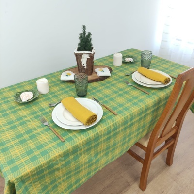 Mesa posta com toalha xadrez verde e amarela, pratos e copos, guardanapos amarelos e vaso com árvore pequena.