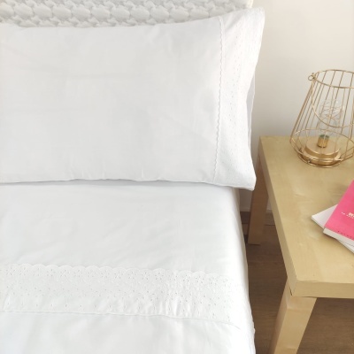 Lençol e fronha brancos com bordado ao lado de mesa de cabeceira com candeeiro e livros