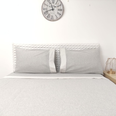 Cama com roupa de cama cinza e branca, cabeceira branca texturada e relógio de parede com números romanos