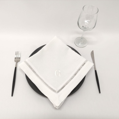 Conjunto de mesa com prato negro, guardanapo branco bordado com letra G, talheres e copo de vidro
