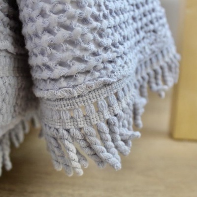 Tecido cinzento claro com textura e franjas torcidas ao lado de móvel de madeira clara