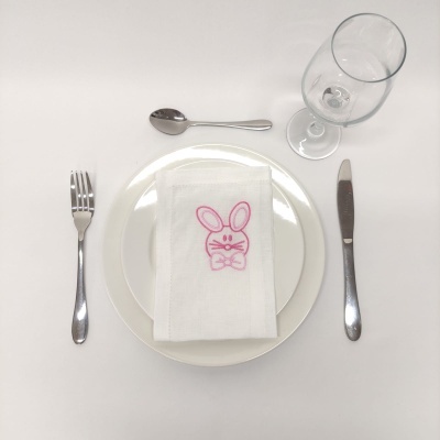 Mesa posta com prato branco, guardanapo branco com bordado de coelho rosa, talheres e copo de vidro