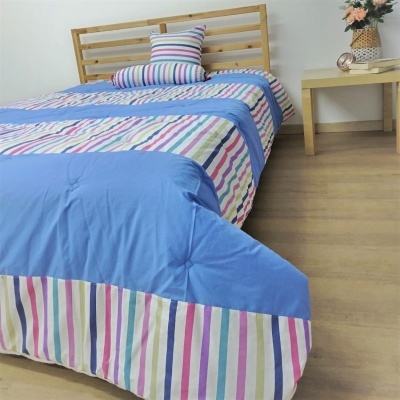 Colcha de cama azul com riscas multicoloridas, almofada e cama de madeira clara ao lado de mesa com flores.