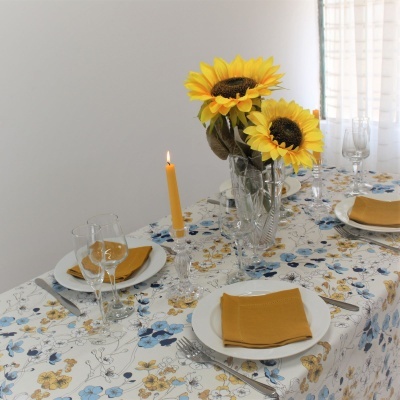 Mesa posta com toalha flores, pratos brancos, guardanapos amarelos, talheres, copos, vaso com girassóis e vela acesa