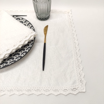 Prato branco com padrão preto, guardanapo branco com renda, colher pequena dourada e preta, copo de vidro texturizado e toalha branca com renda.