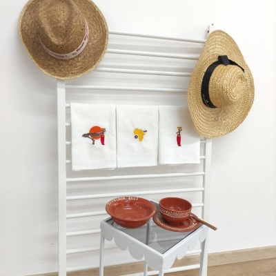 Suportes de parede com toalhas bordadas, chapéus de palha, tigelas de cerâmica e mesa branca em ambiente interno
