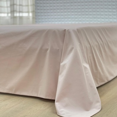 Saia de cama rosa claro com tecido liso sobre cama, chão de madeira clara e cabeceira branca ao fundo