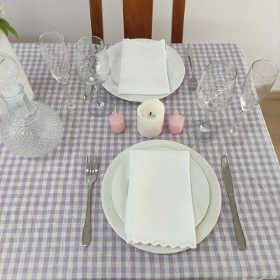 Mesa posta com toalha xadrez lilás e branca, pratos, guardanapos brancos, talheres, copos de vidro e velas no centro