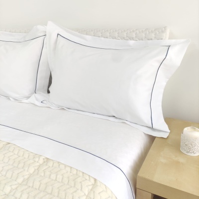 Roupa de cama branca com bordas azuis finas, manta bege texturada e mesa de cabeceira com vela branca