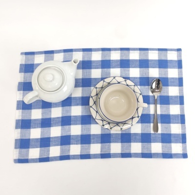 Conjunto de chá com bule branco, chávena bege e azul, colher de metal e toalha aos quadrados azul e branco.