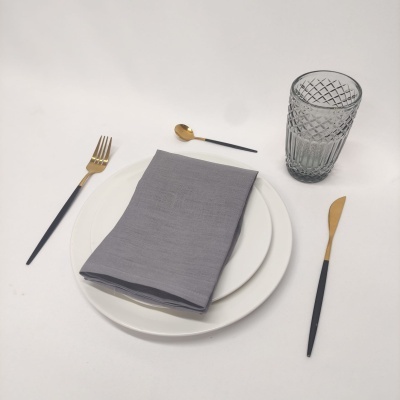 Conjunto de mesa com pratos brancos, guardanapo cinzento, talheres dourados e pretos e copo de vidro cinza numa mesa branca.