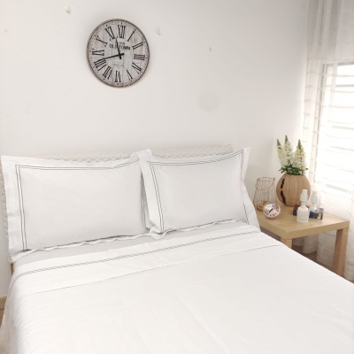 Cama branca com fronhas e lençóis, relógio de parede num espaço iluminado e decorado.