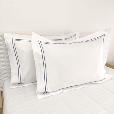 Duas almofadas brancas com bordado azul escuro, sobre cama com lençol branco e cabeceira texturizada