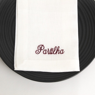 Guardanapo branco com bordado 'Partilha' sobre prato preto com textura circular