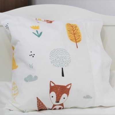 Almofada branca com padrão infantil de raposa, folhas e árvore sobre cama branca junto a berço branco