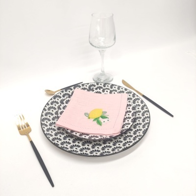 Conjunto de jantar com prato decorado a preto e branco, guardanapo rosa com limão bordado, talheres dourados e pretos e taça de vidro.