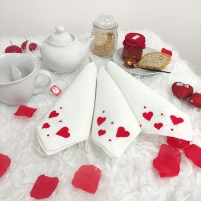 Guardanapos brancos decorados com corações vermelhos, jarro de vidro com bolachas, frasco de compota, chávena e chaleira brancos num cenário com pétalas e chocolates em forma de coração