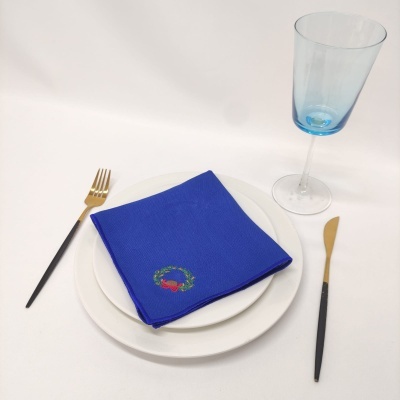 Conjunto de mesa com prato branco, guardanapo azul, talheres com cabo preto e dourado, copo azul sobre superfície branca