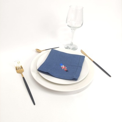 Conjunto de jantar com pratos brancos, guardanapo azul bordado, talheres pretos e dourados, copo de vidro