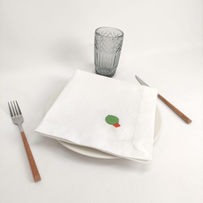 Guardanapo branco bordado com árvore sobre prato branco com talheres de madeira e copo cinzento.