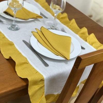 Mesa de madeira com toalha branca e borda amarela, pratos brancos, guardanapos amarelos e copos de vidro