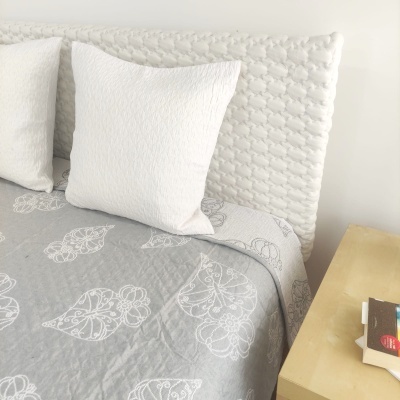 Almofada branca e capa de edredão cinzenta com padrão de corações brancos ao lado de cabeceira acolchoada branca e mesa amarela com livros