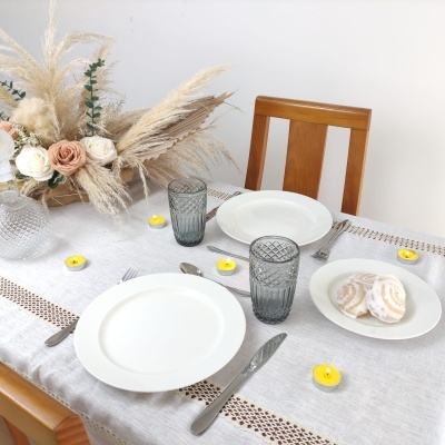 Mesa posta com pratos brancos, copos cinza, velas amarelas e arranjo floral seco em cadeira de madeira clara