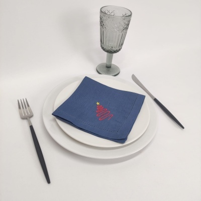 Conjunto de prato branco, garfo e faca com cabos pretos, copo de vidro cinzento com padrão, guardanapo azul com árvore de Natal bordada