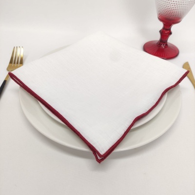 Guardanapo branco com borda vermelha em prato branco com talheres dourados e taça vermelha