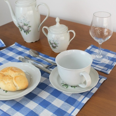 conjunto de chá em porcelana branca com decoração de flores, pão num prato, copo de vidro e talheres sobre toalha azul e branca