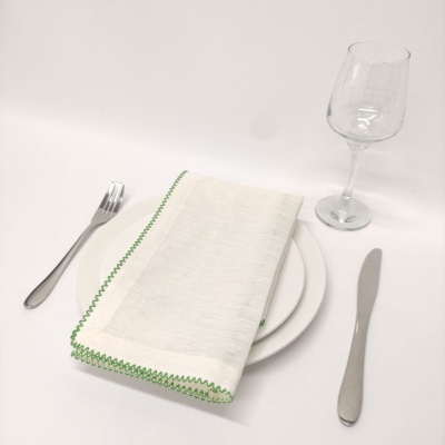 Conjunto de mesa com prato branco, guardanapo branco com borda verde, talheres de metal e copo de vinho