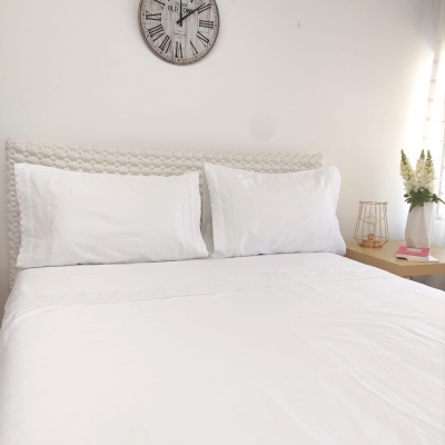 Cama com lençóis e fronhas brancos e cabeceira bege matelassê numa sala com mesa de cabeceira amarela e relógio de parede