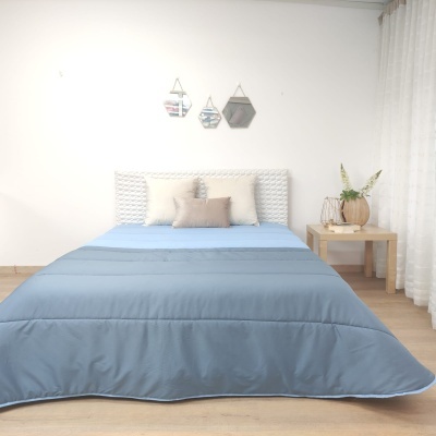 Cama arrumada com roupa azul e almofadas, mesa de cabeceira com vaso e flores, parede branca com espelhos geométricos
