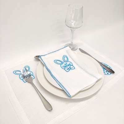 Mesa posta com guarda-napo branco bordado com coelho azul, talheres, prato branco e copo de vidro
