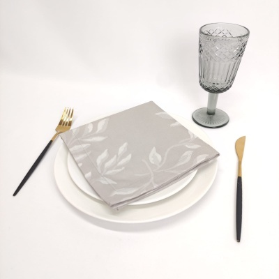 Conjunto de mesa com prato branco, guardanapo cinzento com padrão floral, talheres com cabo preto e dourado, copo de vidro cinzento com textura