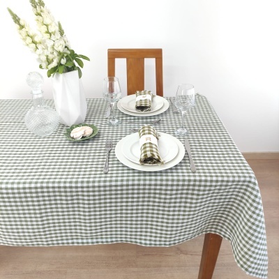 Mesa posta com toalha xadrez verde e branco, pratos brancos, guardanapos combinando, copos de vidro, vaso com flores e decanter em vidro.
