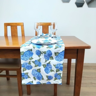 Caminho de mesa branco com flores azuis sobre mesa de madeira clara com copos e prato