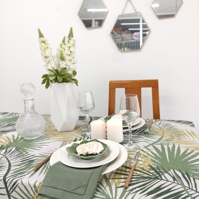 Mesa de jantar com toalha de folhas, plato e guardanapo verde, vaso branco com flores brancas, velas acesas, copos, cadeira de madeira e espelhos hexagonais na parede