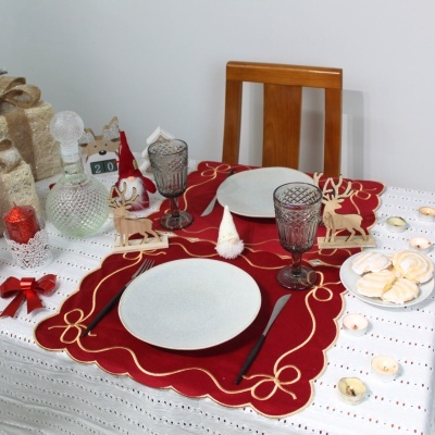 Mesa de Natal decorada com toalha branca, jogos americanos vermelhos bordados, pratos brancos, copos cinza, talheres pretos e enfeites natalícios