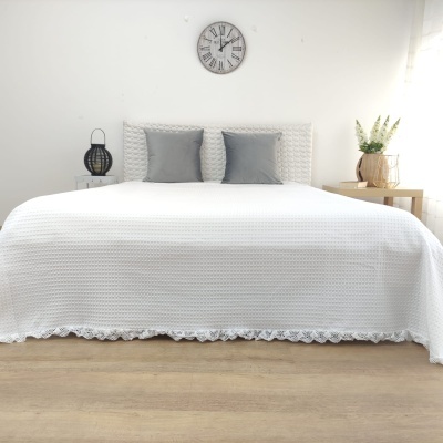 Cama arrumada com colcha branca texturada, duas almofadas cinzentas e cabeceira branca sob relógio de parede, com candeeiro e planta em mesas laterais.