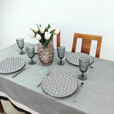 Mesa posta com toalha cinza, pratos pretos e brancos, copos cinza, talheres de aço, vaso rosa com flores brancas e cadeiras de madeira.