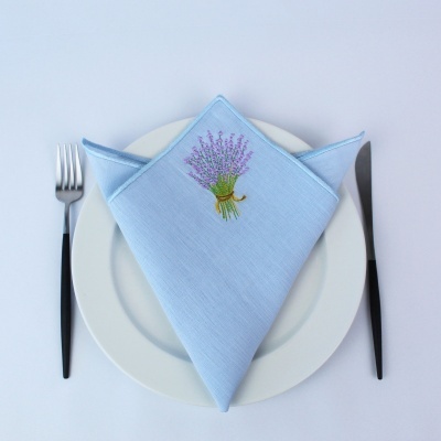 Guardanapo azul bordado com ramo de lavanda sobre prato branco e talheres pretos.